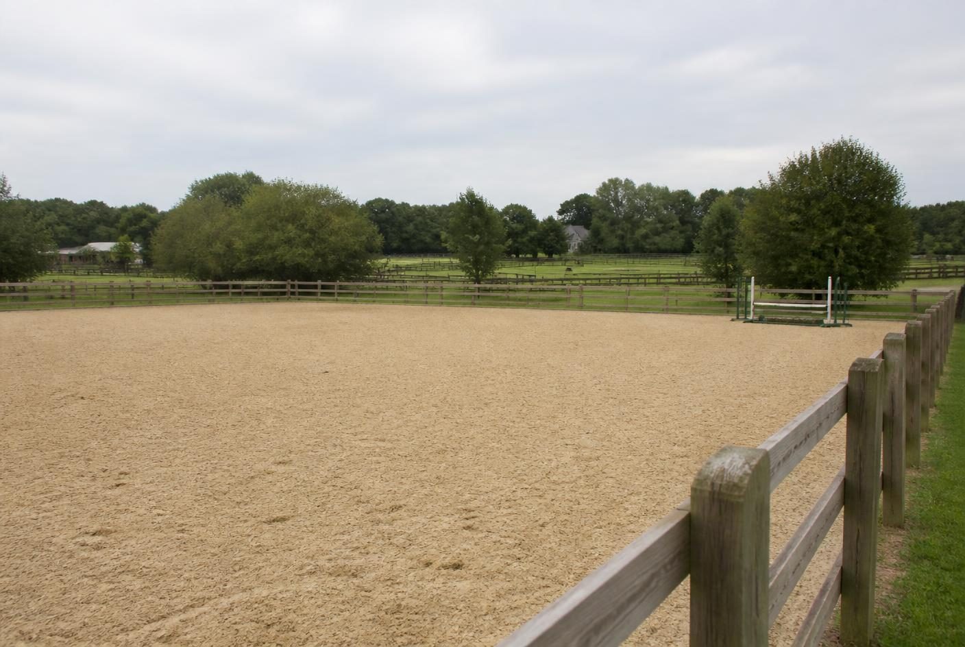 Outdoor sand riding arena with post-and-rail surround and paddocks beyond